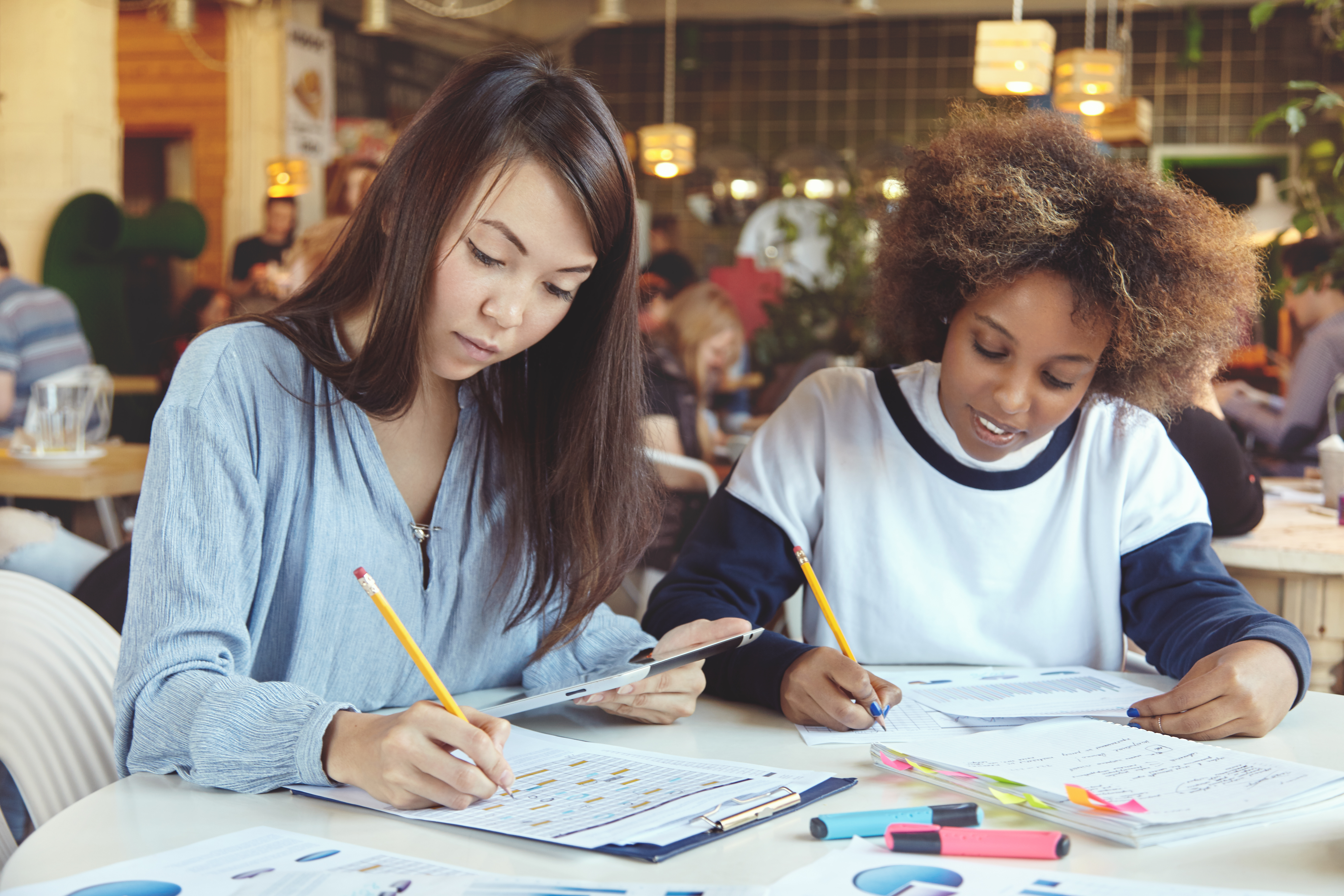 Two female partners working on assignment sitting at table with notebooks and digital tablet, writing down plans and ideas for their start up project. Asian girl holding touch pad and making notes
